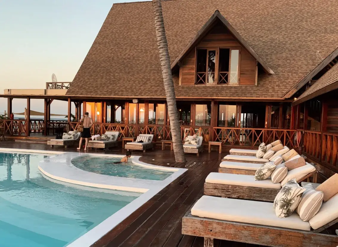 Piscina y tumbonas de un hotel con edificio de madera, al atardecer